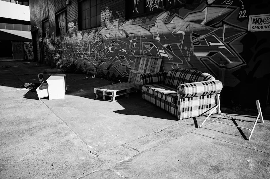 A collection of discarded household furniture and waste materials arranged outside in an urban area, featuring a plaid upholstered sofa placed on the pavement with its legs detached and leaning agains