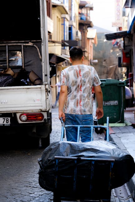 A collection of discarded household furniture and waste materials arranged outside in an urban area, featuring a plaid upholstered sofa placed on the pavement with its legs detached and leaning agains