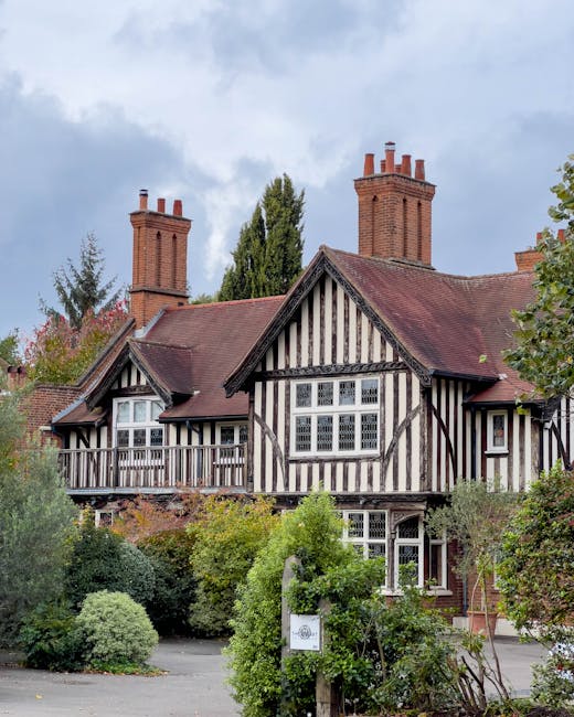 A natural outdoor scene featuring a group of five deer with brown and white coats grazing on a grassy area surrounded by sparse reeds. Behind the deer, there are several leafless trees with dark branches set against a sky with dark clouds and patches of light, indicating late afternoon or early evening. In the background, a distant city skyline with tall buildings illuminated by warm light is visible, contrasting with the natural foreground. The scene appears to be part of a home relocation or moving context, with the focus on the outdoor environment near Richmond Park, as detailed on the page about moving into TW10 homes near Richmond Park by Man with Van Petersham. The image captures the transition between natural and urban landscapes, suitable for supporting content related to house removals, transport, or packing processes in a suburban or park-adjacent setting.