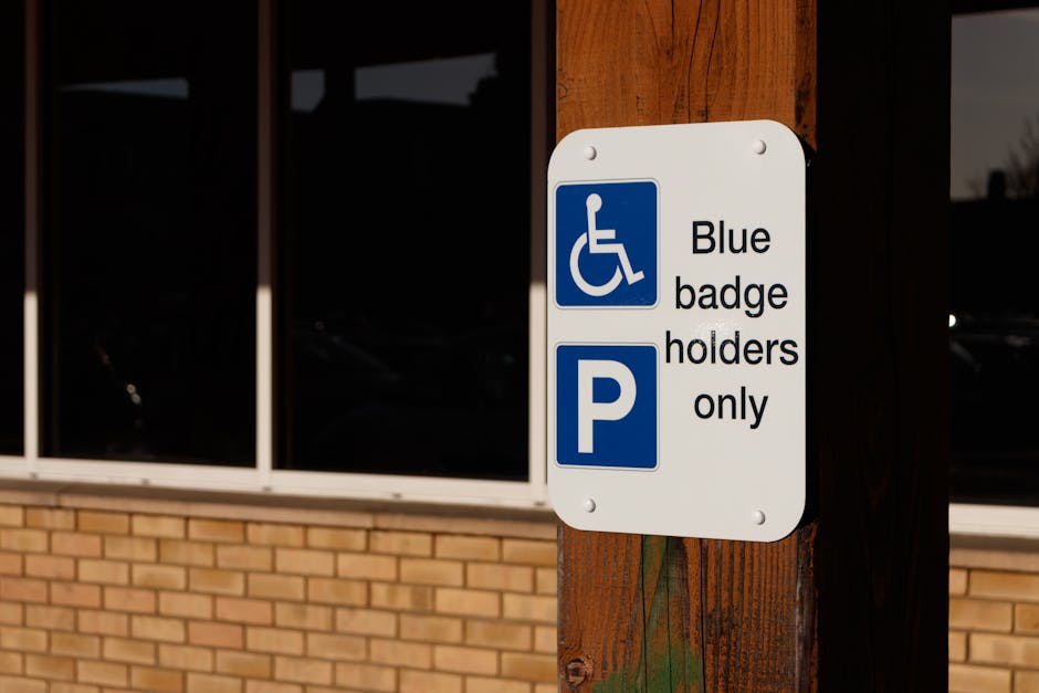 A white rectangular parking sign mounted on a wooden post in front of a brick building with large window panes. The sign includes a blue wheelchair symbol on the top left, indicating accessibility, followed by black text that reads 'Blue badge holders only'. Beneath this, a larger blue parking symbol with a white 'P' is displayed. The sign is positioned at the entrance of a property, suggesting designated parking for disabled badge holders, which may impact vehicle access during a home relocation or furniture transport process. The surrounding environment is well-lit, and the sign's placement signifies parking restrictions or allowances relevant to house removals and moving services provided by Man with Van Petersham, supporting efficient logistics during the moving process in the TW10 area.