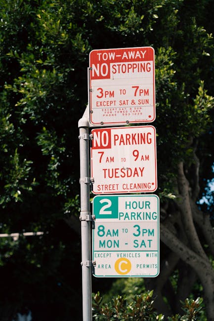 A close-up view of a metal street signpost featuring three rectangular signs stacked vertically. The top sign is red and displays parking restrictions, including a tow-away notice, with white text indicating no stopping from 3pm to 7pm and no parking from 7am to 9am on Tuesdays, with street cleaning times also noted. The middle sign, also red, indicates no parking from 8am to 3pm, Monday through Saturday. The bottom sign has a white background with green and yellow colors, indicating a two-hour parking limit from 8am to 3pm, Monday to Saturday, with a small icon and text referencing permitted vehicles with an area C permit. The signpost is situated on a paved pavement, with dense greenery and trees in the background, typical of an urban residential area in Petersham, London, relevant to house removals and moving logistics in TW10.