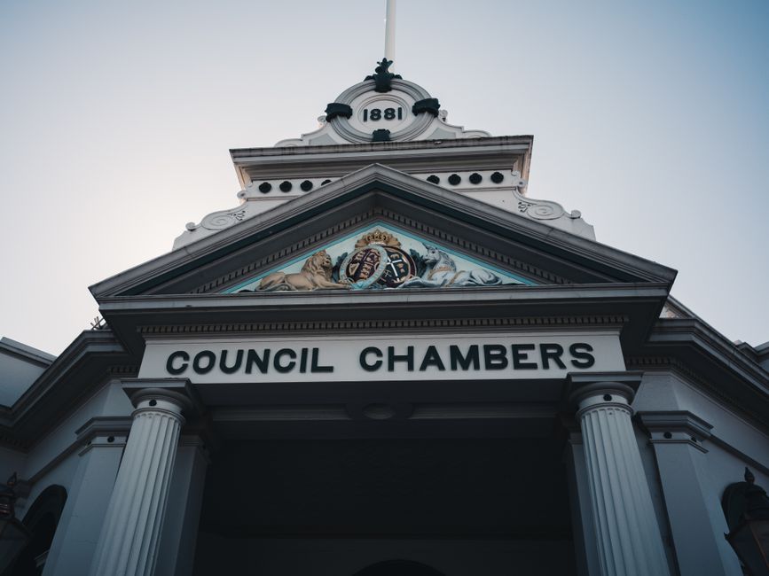A low-angle photograph of the front facade of a council chamber building, displaying classical architectural design with white columns supporting a pediment that features intricate painted details and a coat of arms at the center. The date '1881' is prominently displayed at the top of the structure. The clear sky in the background provides natural lighting that highlights the detailed molding and decorative elements on the building's exterior. Inside the context of house removals and relocation services, this image relates to the process of obtaining necessary permits for property access, such as those explained on the Richmond Council permits page by Man with Van Petersham, a professional removals company offering services in the Petersham area for furniture transport and home relocation.