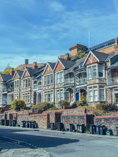 Photograph of a row of Victorian-style terraced houses with ornate white ironwork on balconies and decorative trim around large bay windows. The houses are built with red brick and have pointed gable roofs, some with small garden areas enclosed by brick walls, and are set along a street with black wheelie bins lined up at the curb. The sky above is clear with a few wispy clouds, and the scene depicts a residential area suitable for house removals and relocation services. Man with Van Petersham often facilitates property moves in this locality, ensuring efficient furniture transport and packing for home relocations, as illustrated by the urban streetscape.