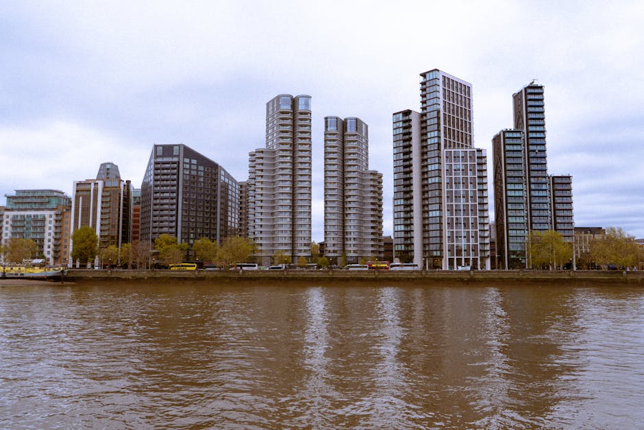 A cityscape view of modern high-rise buildings along a riverbank, with some structures featuring rounded and rectangular architectural designs. The buildings are constructed with glass and concrete materials, reflecting overcast sky conditions. Several trees with green foliage line the promenade area at the water’s edge. In the foreground, the river displays gentle ripples, with a retaining wall or promenade separating the water from the urban area. The scene shows no visible moving vehicles or people, only stationary buildings, and the natural environment bordering a dense cluster of contemporary residential and commercial towers, creating a backdrop relevant to house removals and urban furniture transport. The image captures the riverside environment typical for relocating residential items, supported by the context of a professional removals service such as Man with Van Petersham, which offers packing and moving solutions within this city setting.