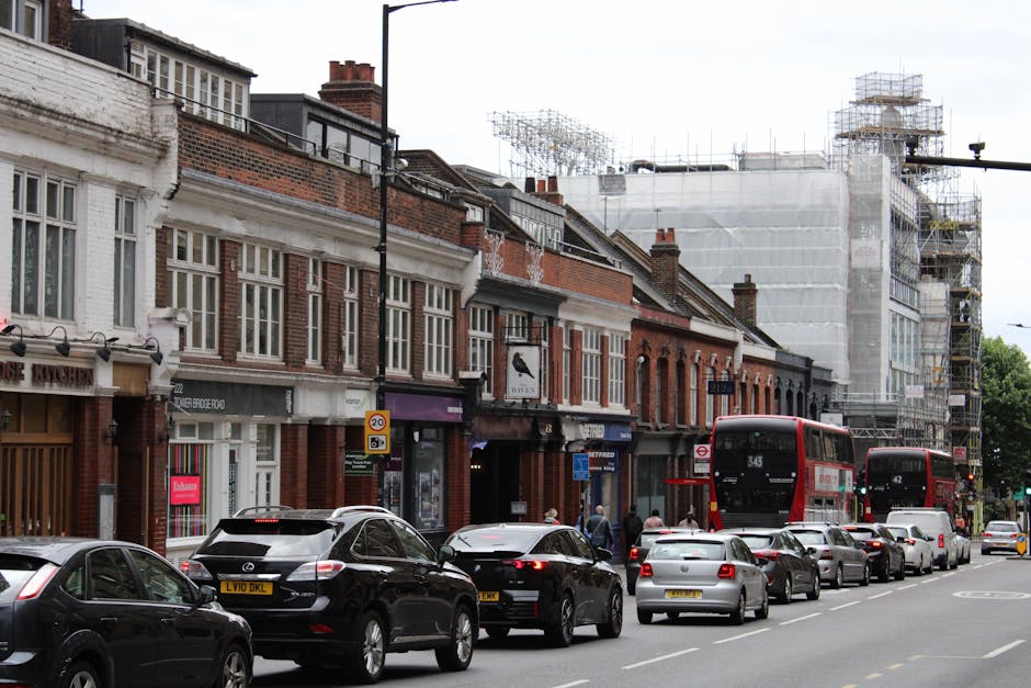 A busy urban street scene featuring a row of brick-fronted shops and residential buildings, with scaffolding and construction work visible on the upper floors of a building in the background. Parked cars, including sedans and an SUV, line the curb, while three red double-decker buses move along the street. Pedestrians are seen walking on the sidewalk, some near the shop entrances. The scene is set during daytime with overcast lighting. This image illustrates a typical street environment during a home relocation process, likely involving furniture transport or packing and moving activities associated with professional removals by Man with Van Petersham. The surroundings show a mixture of commercial and residential properties, with vehicles possibly being loaded or unloaded nearby, emphasizing the importance of managing street parking and traffic during relocations in Petersham (TW10), as addressed on the company’s webpage about skip permits and fines.