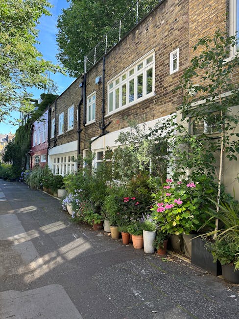 The image depicts the exterior of a residential building with a brick facade, situated along a narrow sidewalk. Several windows are visible on the upper and lower floors, with white frames and multiple panes. The lower level features large bay windows, and the building's exterior is partially shaded by sunlight filtering through nearby trees. In front of the building, there is a row of potted plants and flowers, including leafy shrubs, flowering plants in pink, purple, and white, housed in various containers such as terracotta pots, white ceramic pots, and black plastic planters. The plants are densely arranged, creating a lush appearance. To the left, the sidewalk extends along the street, which appears quiet and residential. The image conveys a calm, tidy environment typical of a home undergoing a furniture and belongings relocation, with Man with Van Petersham likely operating in the area for home removals and moving services, supporting efficient packing, packing and moving, and furniture transport processes.