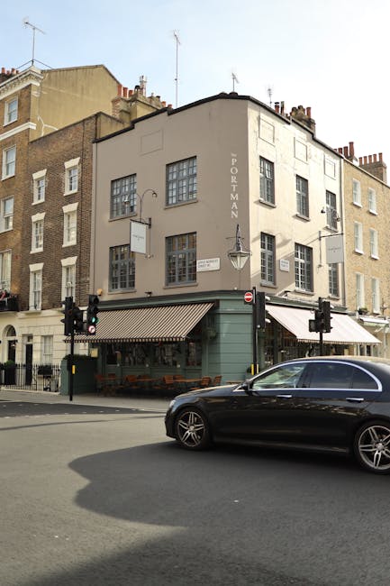 The image shows the exterior of a three-storey building situated at a street corner in Petersham, with a sign indicating 'Portman.' The ground floor features a small café or restaurant with outdoor seating, bordered by a striped awning, and traffic lights are visible at the intersection. In the foreground, a black car is passing by on the street, casting a shadow on the pavement. The building's upper floors have multiple windows, some with small panes, and the overall scene is illuminated by natural daylight. This setting is relevant to house removals and home relocation services, emphasizing urban moving logistics, such as loading furniture and boxes through street access, possibly in relation to small flat moves by [COMPANY_NAME] on this site.