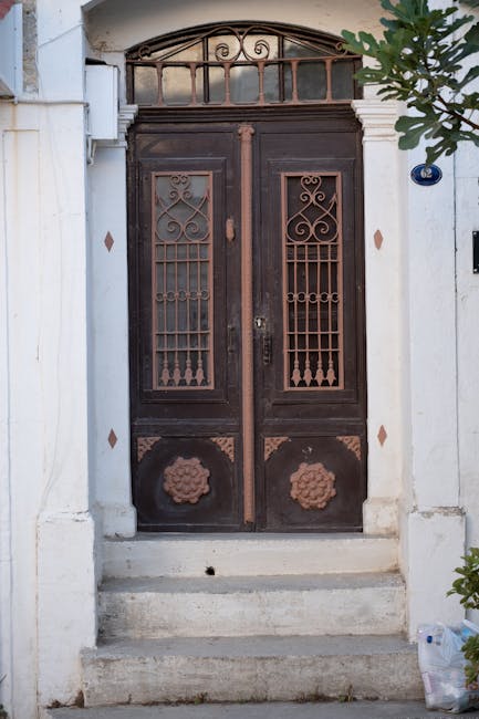 A pair of dark wooden doors with decorative ironwork and floral motifs at the bottom, set within a stone entranceway with a small set of steps leading up to them. The doors are framed by a white exterior wall, featuring a transom window with curved decorative ironwork above. To the right, a green leafy plant partially extends into the frame, and a small blue and white house number plaque is visible on the wall. This scene appears to be the entrance of a residential property, with natural light illuminating the area, suggesting daytime. The image relates to house removals and moving services, highlighting the careful handling of property features during a home relocation. Man with Van Petersham, a professional removals company, may be involved in logistical planning for moving through narrow and delicate entry points like this one.