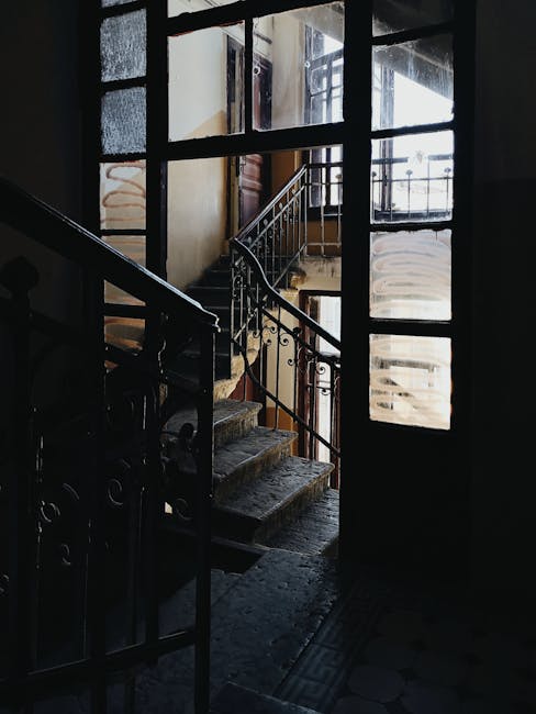 Inside a residential property during a home relocation, a narrow staircase with wooden steps and ornate metal balustrades leads up from a dark hallway illuminated by natural light streaming through large, multi-pane sash windows. The windows show signs of age, with some panes obscured or foggy, and the staircase is positioned adjacent to a tall, glass-paned door. To the side, various packing materials such as cardboard boxes and wrapped antiques are visible, indicating ongoing packing and moving activities as part of furniture transport. The environment appears aged, with textured walls and a worn floor, typical of period homes preparing for a delicate removal process. The lighting highlights the contrast between the dim interior and the bright exterior, emphasizing the careful navigation required to move fragile items through narrow doorways and confined spaces, with professional movers sometimes using trolleys, straps, or blankets, as suggested by the context of [COMPANY_NAME] and their delicate moves service.