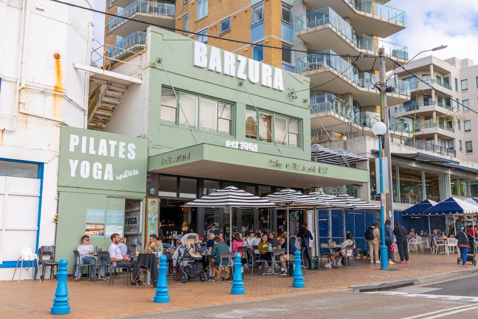 The image shows a busy street scene in front of a two-story restaurant building with a light green facade, featuring large windows and outdoor seating arranged under black-and-white striped umbrellas. Groups of people are seated at tables, some engaging in conversation, others using their phones, with a baby stroller placed nearby. To the left of the restaurant, there is a sign advertising pilates and yoga classes. The sidewalk area is equipped with blue bollards to prevent vehicle encroachment, and pedestrians are walking past the establishment. In the background, a modern multi-story residential building with balconies and glass railings is visible, along with a utility pole and power lines. The scene takes place during daylight hours, with a clear sky overhead. The image exemplifies urban hospitality and the vibrant movement typical of towns like Petersham, where local businesses and community activities intersect, relevant to services such as house and furniture removals facilitated by [COMPANY_NAME] during home relocations or moving logistics in the area.