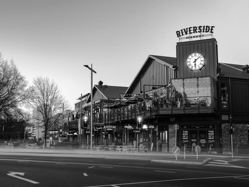 A black-and-white image of a modern building housing Riverside Market, seen from across a wide road with multiple lanes. The building features a prominent clock at the top and a large outdoor balcony area enclosed with glass and railing. On the ground level, there are several storefronts with illuminated signs, including the Riverside Market sign. The scene includes a streetlamp, leafless trees indicating a colder season, and traffic lights showing red signals at the road intersection. This setting represents a commercial area suitable for urban home relocation or furniture transport logistics, with clear views of the building's structure and surrounding environment that could be involved during a house removal or moving process, as handled by [COMPANY_NAME].
