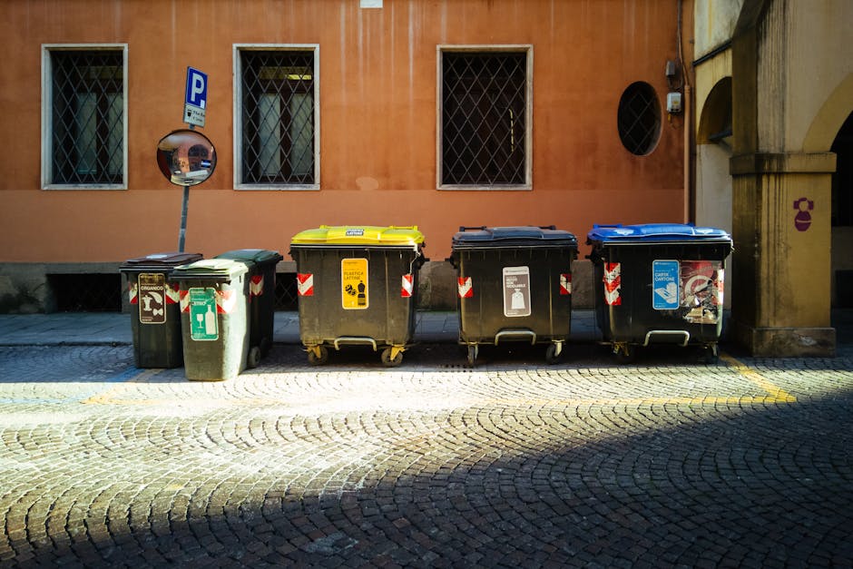 A row of four wheelie waste bins positioned on a cobblestone street in front of a peach-coloured building with three windows, two of which have metal security grilles. The first bin on the left has a green lid and is marked for general waste, with recyclable materials inside visible through the transparent section. The second bin features a yellow lid, designated for electronic waste and contains a cardboard box inside. The third bin has a black lid and is used for general rubbish, with some litter visible inside. The fourth bin has a blue lid and is intended for recyclable plastics, with some plastic packaging attached to its exterior. A convex traffic mirror and a parking sign are mounted on a pole adjacent to the bins, with sunlight casting shadows across the cobblestones, indicating an outdoor setting in a residential area. The image emphasizes personal and commercial waste disposal in relation to house and furniture removals, aligning with property relocation services provided by Man with Van Petersham.