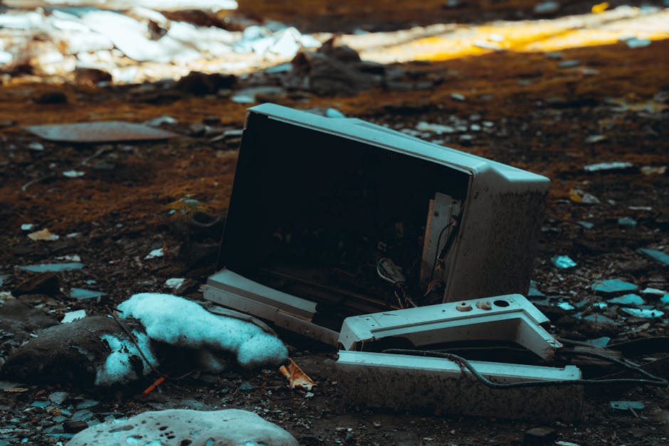 A discarded old microwave oven with a broken door lies on muddy ground outdoors, surrounded by patches of snow and scattered debris such as broken plastic and cardboard. The microwave's internal components are visible through the damaged front, and its exterior is stained and weathered, indicating it has been thrown away or abandoned. The environment appears to be an outdoor dumping area or landfill site, with natural daylight illuminating the scene. This image reflects the disposal process associated with house removals and the proper handling of appliances during moving or waste clearance. Man with Van Petersham, a professional removal company, may assist with the safe disposal or recycling of such household items during furniture transport and logistics in home relocation services, following best practice waste disposal rules for appliances.